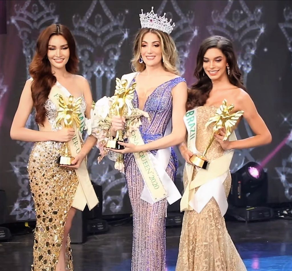 The three finalists of Miss International Queen 2020 on stage in evening gowns, holding trophies